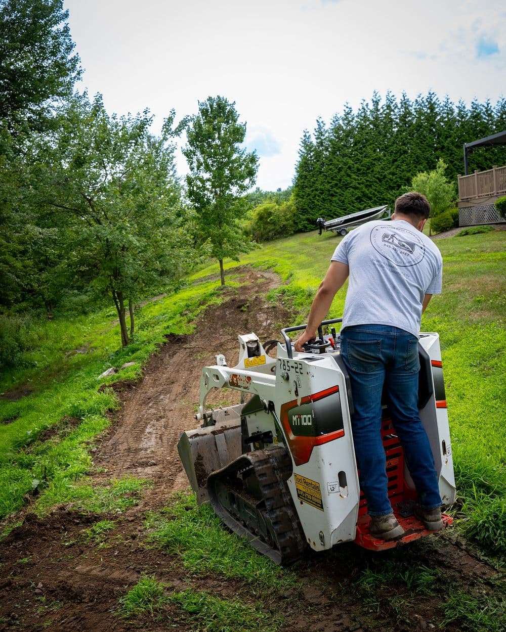Person operating a walk-behind mini skid steer on a landscaped lawn with trees.